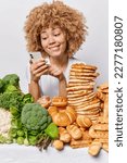 Small photo of Vertical shot of cheerful curly woman checks newsfeed via smartphone orders fodd online keeps to healthy diet refuses eating unhealthy harmful food chooses green vegetables instead isolated on white