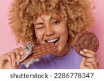 Close up shot of beautiful woman with curly hair bites bar of chocolate holds cookie winks eye has perfect white teeth has addiction to sugar isolated over pink background. Sweet tooth concept
