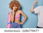 Small photo of Displeased curly haired woman smells something stinky and disgusting looks on sweaty armpit covers nose goes in for sport at gym keeps hand on waist isolated over blue background. Intolerable smell