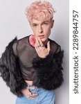 Small photo of Young transgender man touches face looks seriously at camera wears net t shirt black fur coat and jeans tries to seduce someone poses against white background. Lgbt male model in new stylish clothing