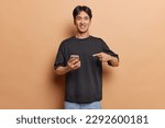 Small photo of Handsome Chinese man smiles gladfully points at his new smartphone shares awesome news with friends dressed in casual black t shirt isolated over brown background asks to check out newsfeed.