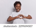 Small photo of Lovely dark skined woman expresses sympathy and admiration makes heart gesture smiles toothily has romantic mood dressed in t shirt isolated over white background. Be my valentine. I love you sign