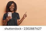 Small photo of Horizontal shot of pretty young African woman smiles gently dressed in casual black t shirt holds mobile phone checks newsfeed points index finger at blank space isolated over brown background