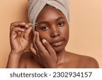 Close up shot of dark skinned woman plucks eyebrows with tweezer removes excess hair stands shirtless looks directly at camera poses with bath towel wrapped on head isolated over brown background