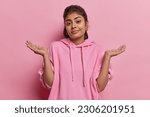 Small photo of Clueless indecisive Indian woman spreads palm shrugs shoulders looks clueless at camera dressed in casual sweatshirt cannot decide or answer your question isolated over pink background faces dilemma