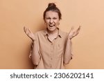 Small photo of Angry fed up young European woman spreads palms and yells annoyed argues with someone wears shirt expresses negative emotions isolated over brown background. How dare you. Furious female model