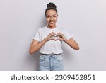 Radiant woman with warm smile shows heart gesture spreads love has heartfelt expression dressed in casual t shirt and jeans stands against white background. Gratitude relationships affection concept