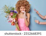 Small photo of Pretty curly haired girl appears sad as she holds bouquet of flowers symbolizing her unfortunate allergy sneezes all time being suggested to take medicine wears pink dress and white lace gloves