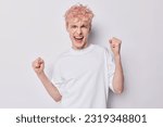 Small photo of Freckled pink haired man clenches his fists in celebration displaying winner gesture exclaims from happiness dressed in casual t shirt isolated over white background. Accomplishment and celebration