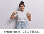 Small photo of Young dark haired woman captures attention with her vibrant energy with contagious smile directs focus to her blank white tshirt using two fingers shows place for your logo or design poses indoor