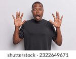 Small photo of Black African man with palms raised in fear his eyes widen as he gasps in astonishment or horror notices something undeniably spooky has reaction capturing essence of surprise wears t shirt.