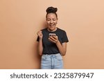 Small photo of Positive Latin woman with hair bun exudes pure amazement and delight with firm fist holds her smartphone capturing thrill of gambling victory exclaims like winner isolated over brown background.