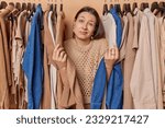 Small photo of Hesitant woman shrugs shoulders feels unaware chooses perfect outfit for occasion wears knitted jumper poses among clothes rail poses clueless lost in quest to find ideal attire for special event