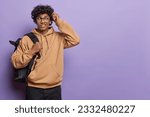 Small photo of Student life concept. Horizontal shot of clueless curly haired man scratches head and looks away with hesitation cannot remember answer on question dressed in brown sweatshirt and black trousers