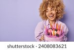 Small photo of Studio shot of glad woman with curly bushy hair holds big tasty cake looks aside happily smiles broadly wears festive clothing celebrates birthday isolated over purple background copy space for text