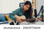 Small photo of Happy asia woman songwriter play acoustic guitar listen song from smartphone think and write notes lyrics song in paper sit in living room at home studio. Music production at home concept.