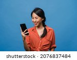 Small photo of Young Asia lady using phone with positive expression, smiles broadly, dressed in casual clothing feeling happiness and stand isolated on blue background. Happy adorable glad woman rejoices success.