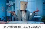 Small photo of Young Asian businesswoman sit on desk wear draw sad mask paper craft bag feel bad mood with fail work project in office at night. Anonymous lady mad and confuse with overwork, Work mental health.