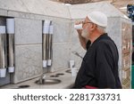 Small photo of Medina, Saudi Arabia - March, 2023 : an old hajj or umrah pilgrim man is drinking zam zam water in the sink of the zam zam water faucet. Water is available free and unlimited.