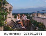 Young couple relaxing outdoors on urban rooftop garden with blooming flowers. Man and woman in casual clothes enjoying sunset and drinking tea or coffee at apartment balcony terrace with city view.