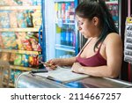 Small photo of latina woman concentrating while calculating the number of sales in a notebook of her grocery store. girl leaning on a glass display case thinking about sales. concept of economy and business