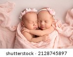 Small photo of Tiny newborn twin girls. A newborn twin sleeps next to his sister. Newborn twin girls on the background of a pink blanket with pink bandages. The girls gently hug and kiss their sister in a cute pose.