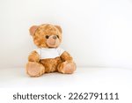 Cuddly brown teddy bear stuffed toy wearing a white t-shirt sitting on white table against white wall, copy space