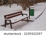 A snowy park bench with a green trash can. 