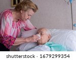 Small photo of Woman attending to her nonagenarian husband, giving him a drink, in hospital bed