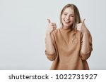 Small photo of Portrait of fair-haired beautiful female student or customer with broad smile, looking at the camera with happy expression, showing thumbs-up with both hands, achieving study goals. Body language