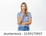 Small photo of Portrait of stylish young woman with blond hair, looking thoughtful, deciding smth, pondering with puckered lips and thinking face expression, standing against white background