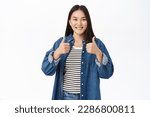 Small photo of Very good. Smiling asian girl shows thumbs up, nod in approval, saying yes, praising smth, standing over white background.