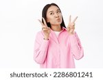 Small photo of Cute and silly asian girl shows peace sign, pucker lips, kissing face, posing in pink shirt over white background.