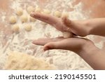 Small photo of Children's hands roll a ball of dough between their palms against the background of already made dough balls, a large piece of dough and a wooden table in flour. The process of making dessert