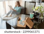 Smartphone is in separate wicker box with inscription digital detox on table. Woman reading book in background. Stop using digital gadgets. Mental and digital detox concept