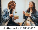 Small photo of Two millennial women female friends sitting outdoors eating takeaway food, laughing and having fun. Food delivery and takeout.