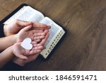 Religious Christian girl praying with her mother indoors. Bible in background. Hands holding white paper heart. Space for text