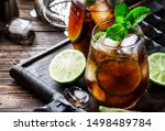 Cuba libre alcohol cocktail with golden rum, lemon juice, cola, lime and ice, dark bar counter background, bar tools, place for text