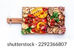 Small photo of Grilled vegetables: red and yellow paprika, zucchini, eggplant, mushrooms, tomatoes and onions served on rustic wooden cutting board, white table background, top view