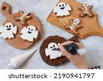 Small photo of Decorating Halloween gingerbreads of ghosts and skeletons with frosting. Girl holds pastry bag with black icing and decorates gingerbread ghost