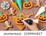 Small photo of Decorating Halloween gingerbreads of pumpkin lantern and skeletons with frosting. Girl holds pastry bag with black icing and decorates gingerbread pumpkin lantern