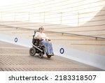 Small photo of a woman with sunglasses sitting in an electric wheelchair going down an urban ramp. Concept of a handicapped accessible city