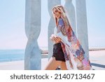 Small photo of Portrait of young beautiful happy smiling blond woman in summertime. Female outdoors wearing colorful smock with marble column background in front of the sea. Summer travel concept.