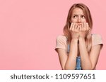 Small photo of Studio shot of anxious nervous female model bites finger nails with puzzlement, dressed casually, stands against pink background with copy space for advertisement. Negative human emotions concept