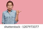 Small photo of Surprised Caucasian young lady has bated breath, dressed in stylish outfit, models against pink background, indicates with thumb at free space, opens mouth widely. Advertisement and shock concept