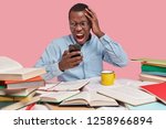Small photo of Irked angered dark skinned young man looks with discontent peevish facial expression at cell phone, dressed in formal shirt, keeps hand on head, surrounded with many books, recieves bad message