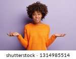 Isolated shot of confused beautiful woman with dark skin, curly haircut, spreads hands sideways, smirks face, feels doubt while makes choice, dressed in casual jumper, isolated over purple wall
