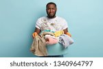 Small photo of Sad pity African American husband overloaded with housework, wears dirty clothes and gloves has duties to wash linen, cries from depression and puzzlement, stands over blue background. Too much work