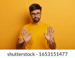 Slow down. Serious looking displeased bearded man shows stop gesture, asks to hold horses, keeps palms towards camera, says take it easy and control your behaviour, stands against yellow background