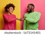 Positive dark skinned young woman and man bump fists, agree to be one team, look happily at each other, celebrates completed task, wear pink and green clothes, pose indoor, have successful deal